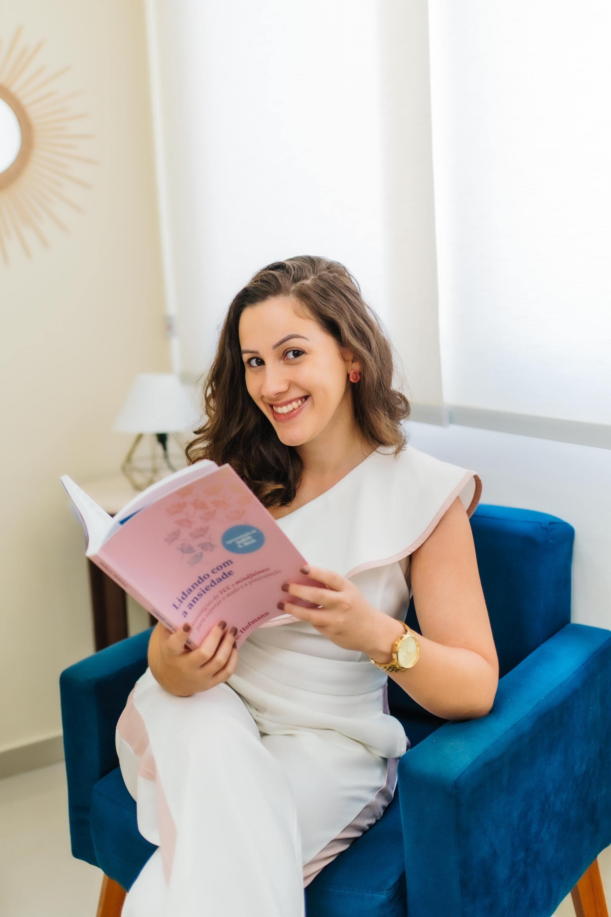 Myllena Barbosa, psicóloga, sentada em uma poltrona azul com um livro sobre ansiedade